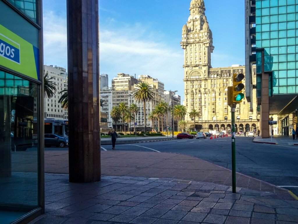 Ampla rua da cidade com palmeiras e uma alta torre histórica ornamentada entre edifícios modernos de vidro Catedral Metropolitana de Montevideo