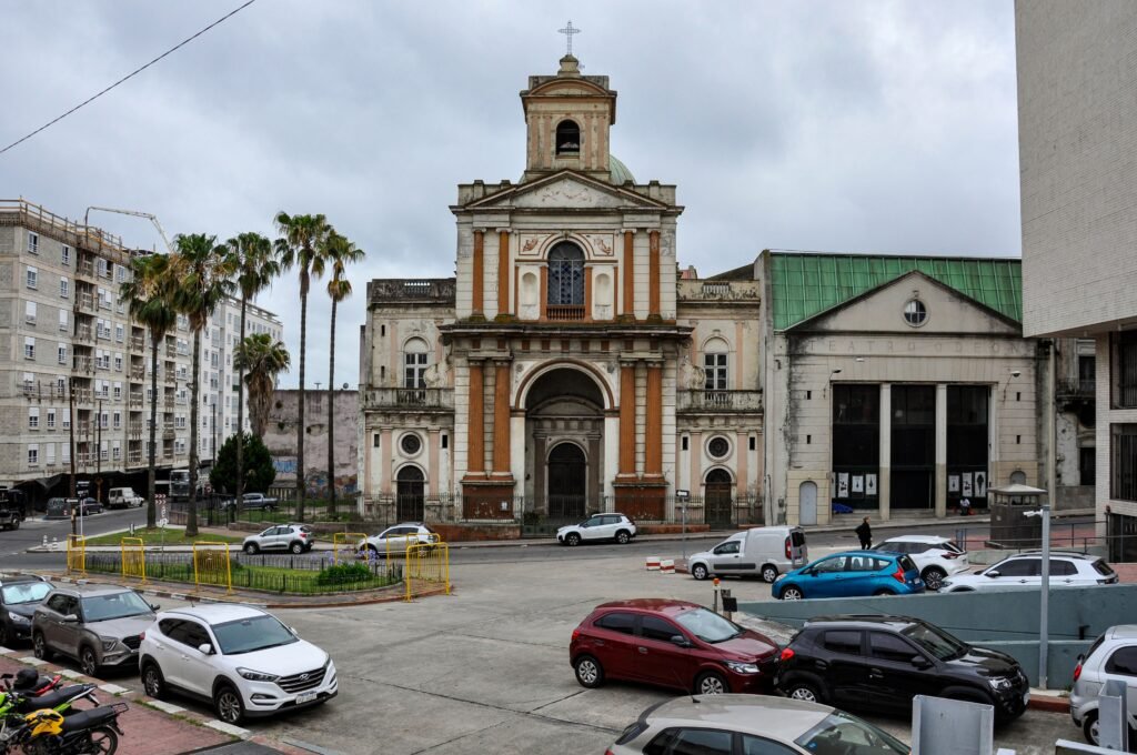Vista frontal de uma igreja histórica desgastada pelo tempo, com campanário e cruz no topo, situada em uma rua movimentada da cidade, com palmeiras e carros estacionados nas proximidades Catedral Metropolitana de Montevideo