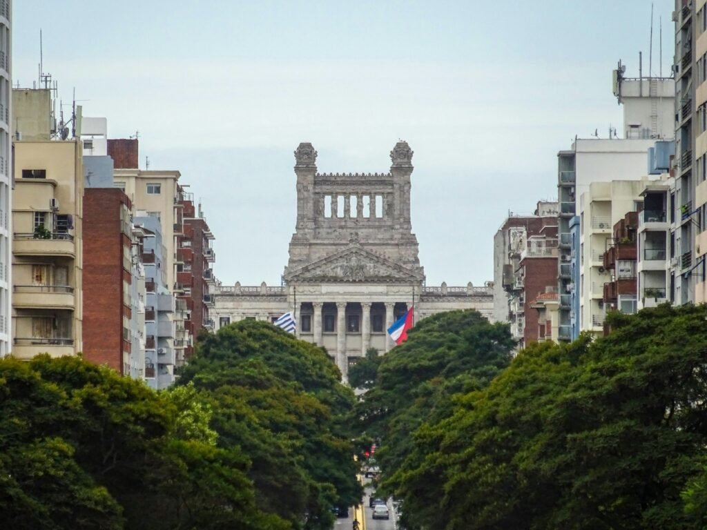 Uma avenida arborizada, ladeada por edifícios de média altura, conduz a um grandioso edifício histórico de pedra com torres ornamentadas ao fundo; em primeiro plano, um exuberante parque verde Catedral Metropolitana de Montevideo
