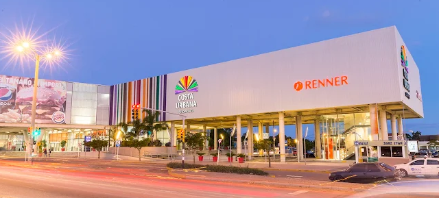 Fachada iluminada do Costa Urbana Shopping ao entardecer com a loja Renner