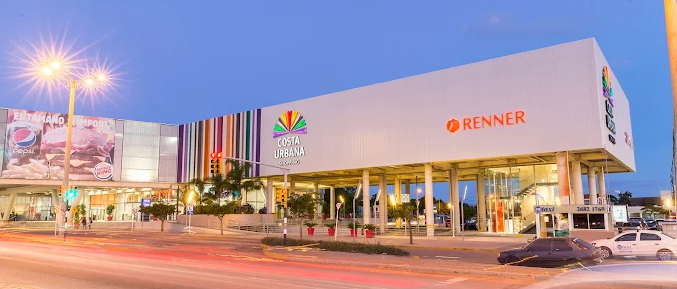 Fachada iluminada do Costa Urbana Shopping ao entardecer com a loja Renner