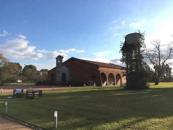 Edifício de tijolos com arcos em um parque gramado, com uma pequena torre e uma caixa-d'água coberta por hera à direita, sob um céu azul.