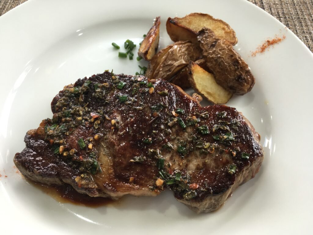 Bife selado coberto com ervas picadas em um prato, acompanhado de fatias de batata assada.