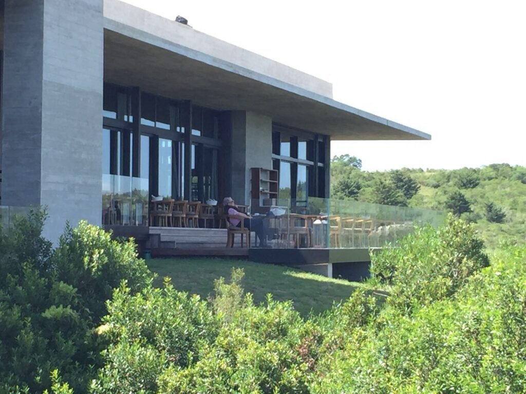 Casa moderna com paredes de vidro, deck sombreado e uma fileira de cadeiras; uma pessoa está sentada e relaxando no pátio.