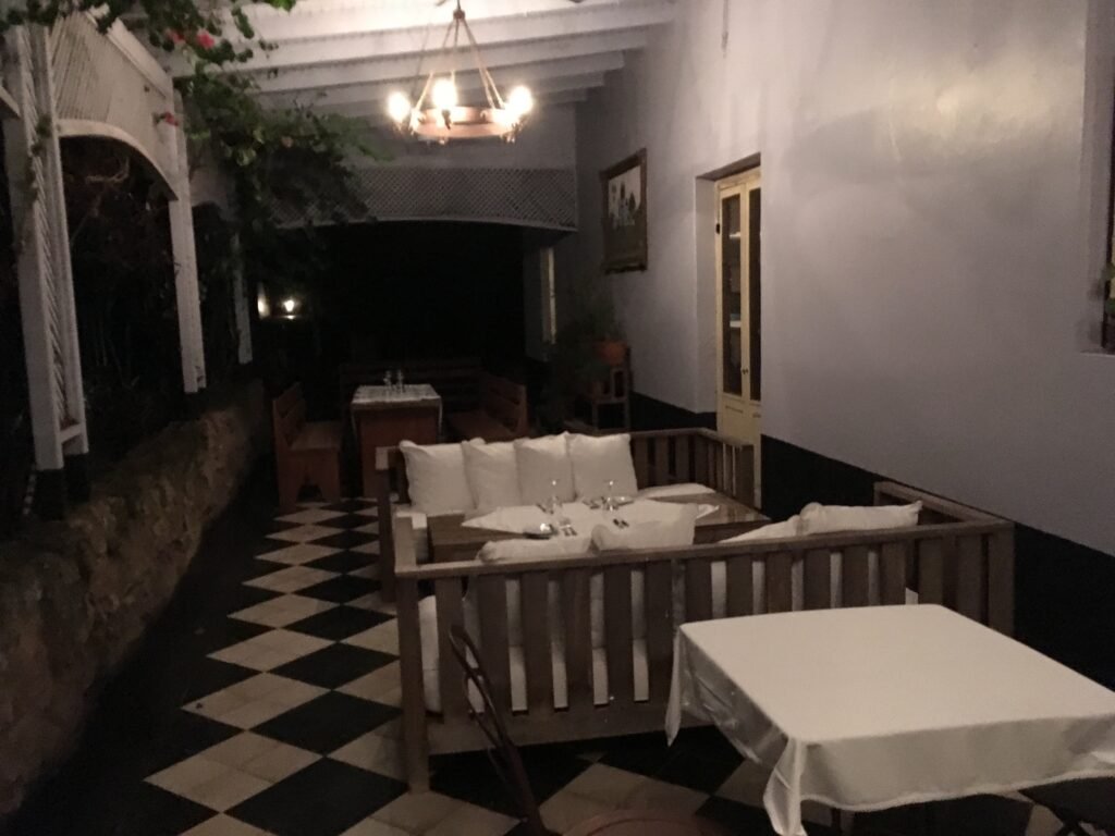 Área de refeições ao ar livre à noite, com assentos de madeira e almofadas brancas, piso xadrez e um lustre suspenso.
