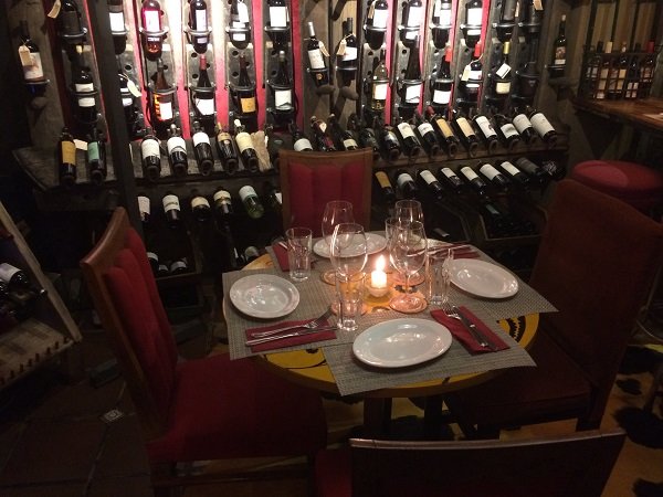 Mesa posta para quatro pessoas, com velas, taças e pratos, tendo como cenário uma adega com prateleiras de vinho que cobrem toda a parede.