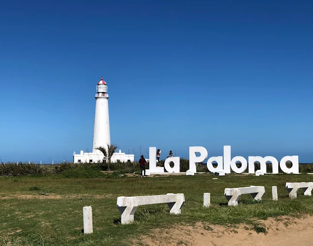 Farol de La Paloma branco ao lado do letreiro La Paloma sob céu azul