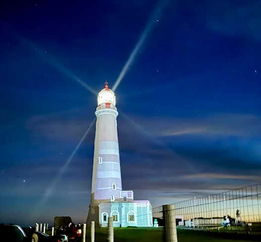Farol de La Paloma iluminado à noite com feixes de luz contra o céu estrelado