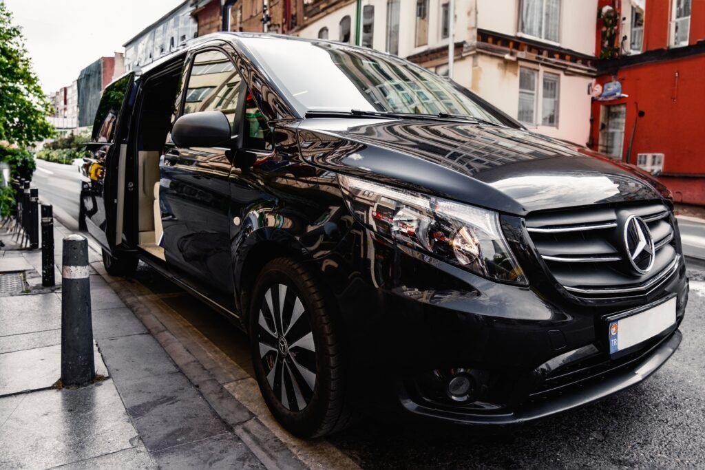 Van Mercedes-Benz preta com a porta lateral aberta, estacionada em uma rua urbana estreita; sua superfície brilhante reflete os prédios e o céu.