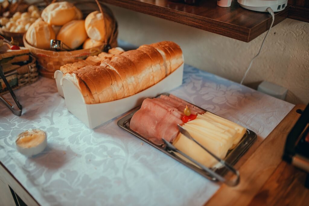 Pão de forma fatiado em uma forma branca, sobre uma mesa de buffet, com fatias de presunto e queijo por perto.