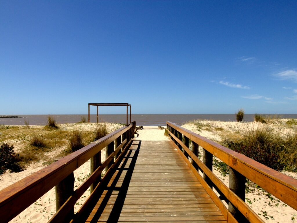 Calçadão de madeira que leva a uma praia sob um céu azul claro, com o mar no horizonte Letreiro de Montevideo em Pocitos