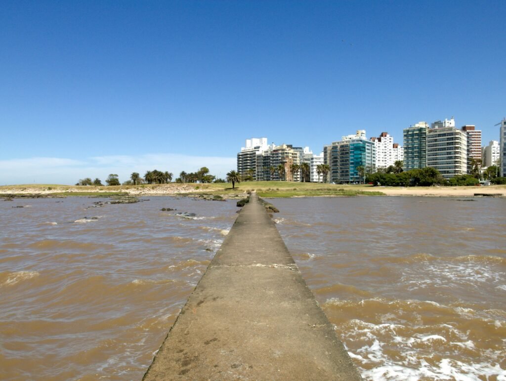 Cais de concreto que se estende pelas águas marrons em direção ao horizonte da cidade, com seus modernos arranha-céus, sob um céu azul límpido Letreiro de Montevideo em Pocitos