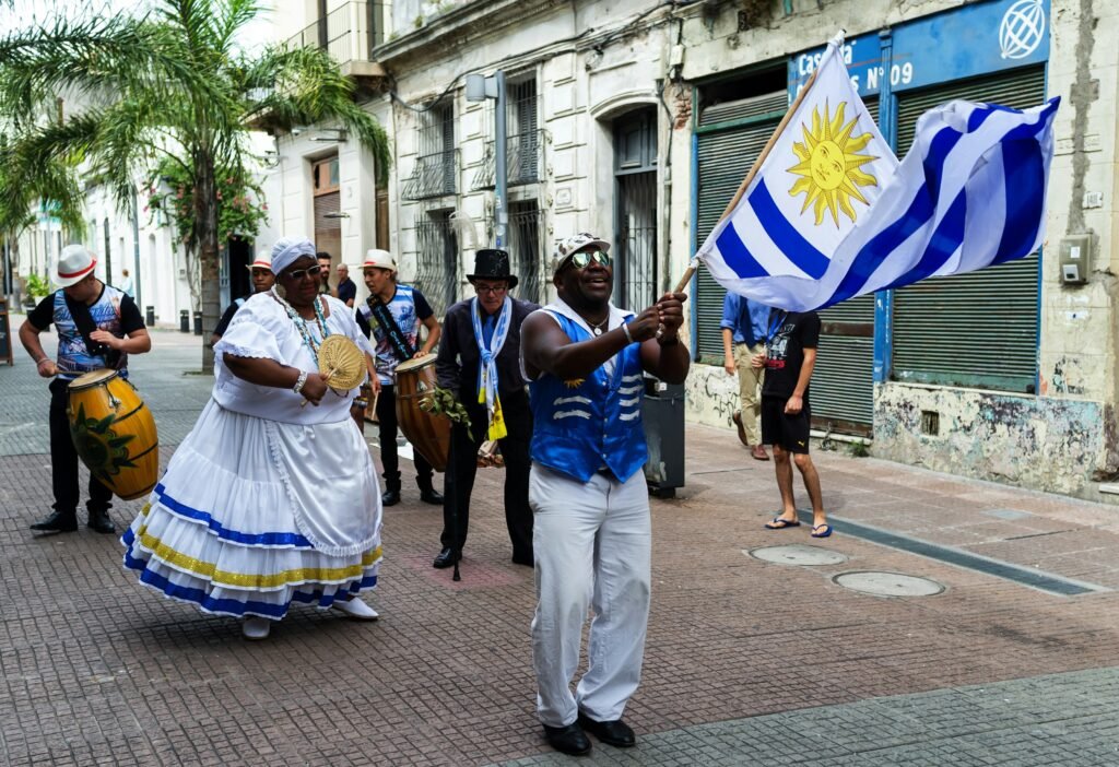 Músicos marcham por uma rua; uma mulher de vestido branco com detalhes azuis toca um leque enquanto outros tocam tambores e um porta-bandeira segura uma bandeira do Uruguai Letreiro de Montevideo em Pocitos