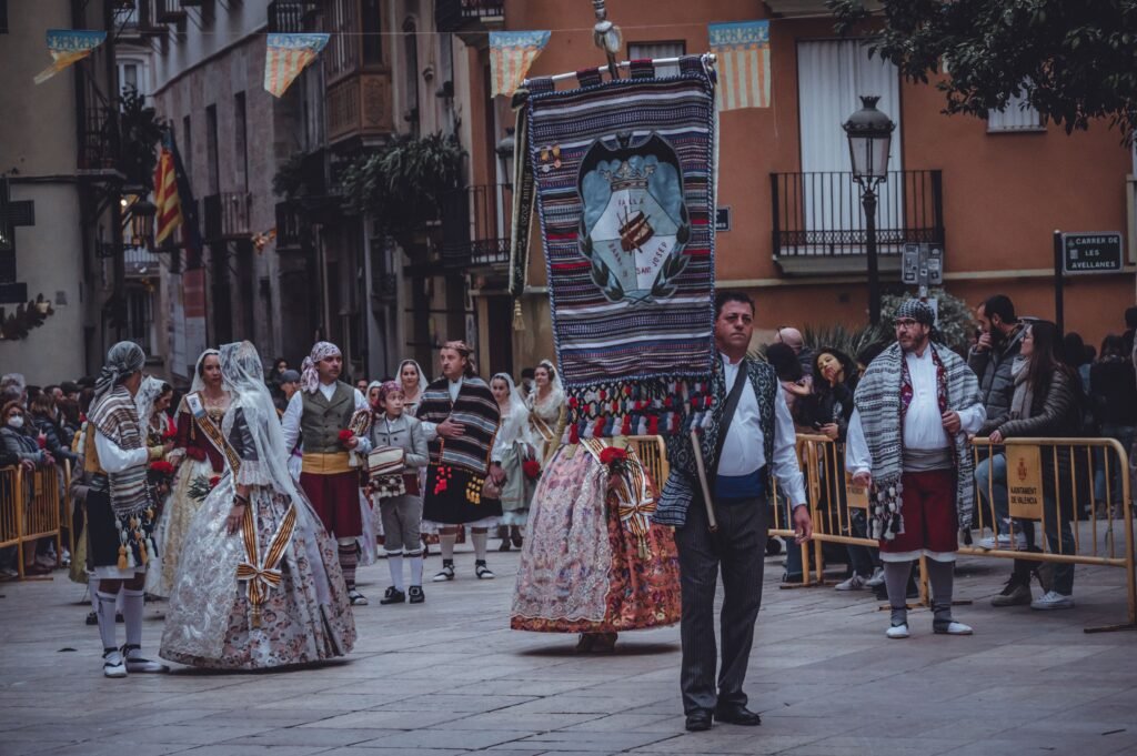 Uma multidão vestida com trajes tradicionais desfila por uma rua da cidade, carregando um grande estandarte cerimonial tecido no centro do palco Museu Espanhol em Colônia do Sacramento
