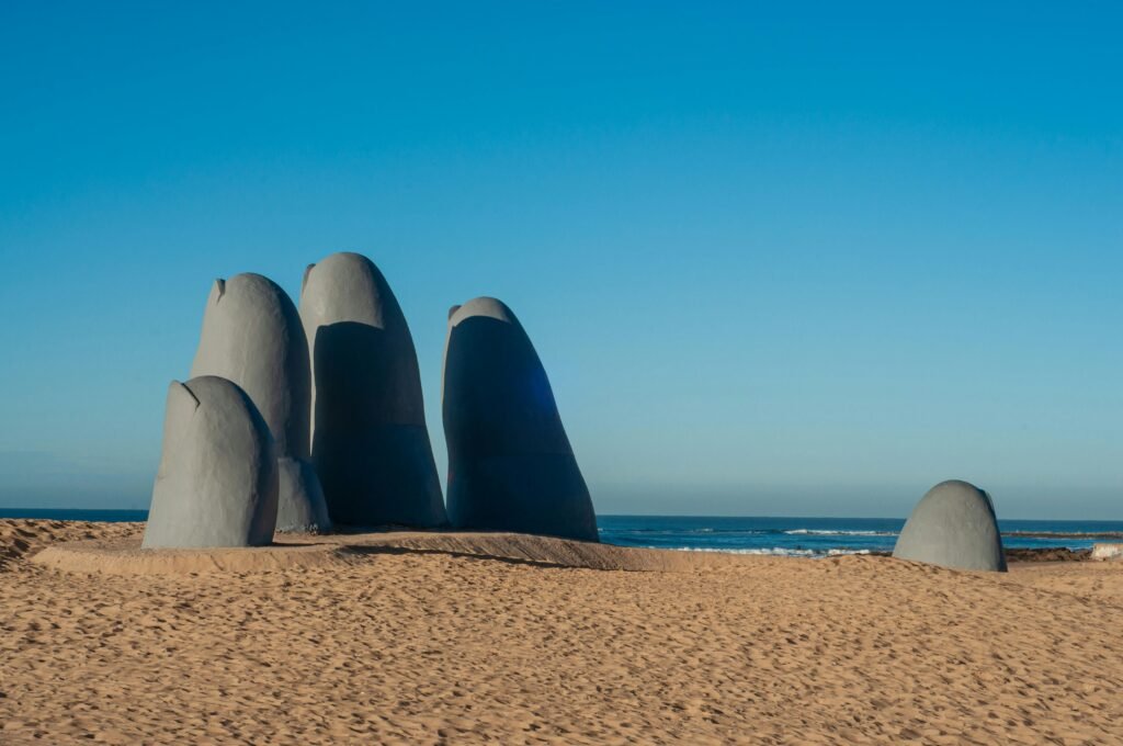 Conjunto de grandes esculturas de pedra arredondadas em forma de dedos, emergindo de uma praia arenosa contra um céu azul claro