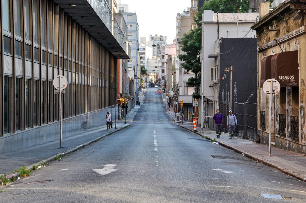 Rua urbana vista de uma leve inclinação, com uma fachada de vidro alta à esquerda e prédios cobertos de grafite à direita, e alguns pedestres à frente.