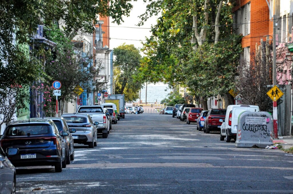 Rua arborizada com carros estacionados em ambos os lados e um vislumbre da água ao final da via.