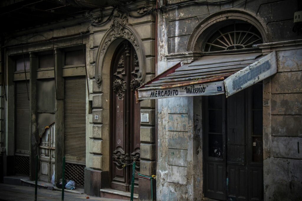 Prédio urbano antigo com porta de entrada em arco decorativa, paredes desgastadas e toldo de fachada esfarrapado com a inscrição 'Mercadito Milenio'.
