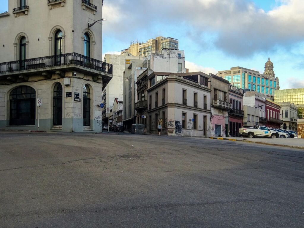 Esquina da cidade com edifício histórico bege à esquerda, varanda de ferro, grafites nas paredes inferiores e edifícios modernos ao longe sob um céu azul com nuvens.