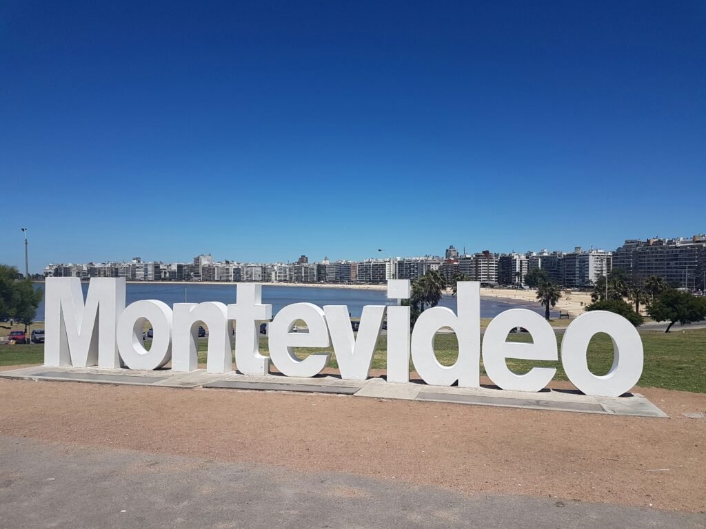Grande placa branca de Montevideo à beira-mar, com o horizonte da cidade e o céu azul ao fundo.