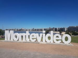 Grande placa branca de Montevideo à beira-mar, com o horizonte da cidade e o céu azul ao fundo.