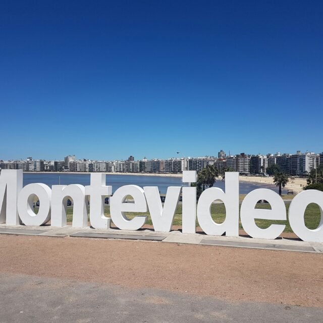 Grande placa branca de Montevideo à beira-mar, com o horizonte da cidade e o céu azul ao fundo.