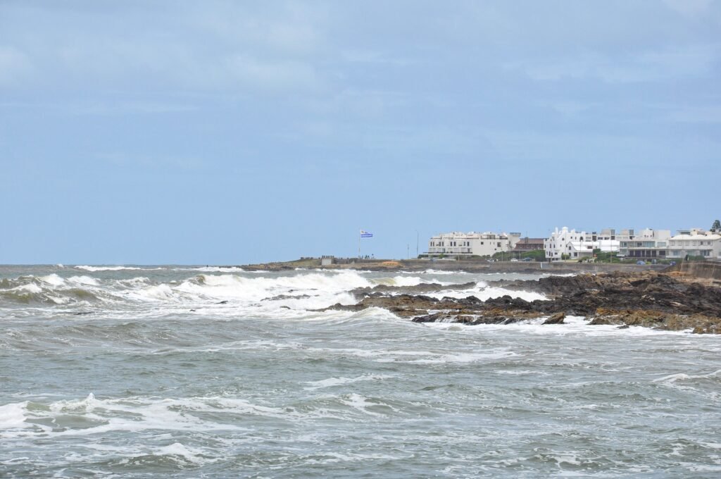 Paisagem costeira com costa rochosa, ondas quebrando perto da margem e construções brancas ao longo do litoral, com uma bandeira ao longe.