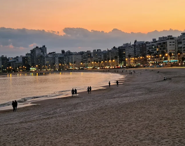 Pessoas caminhando na Praia de Pocitos ao pôr do sol com as luzes da orla acesas