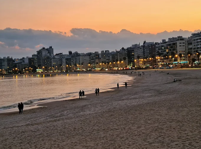 Pessoas caminhando na Praia de Pocitos ao pôr do sol com as luzes da orla acesas