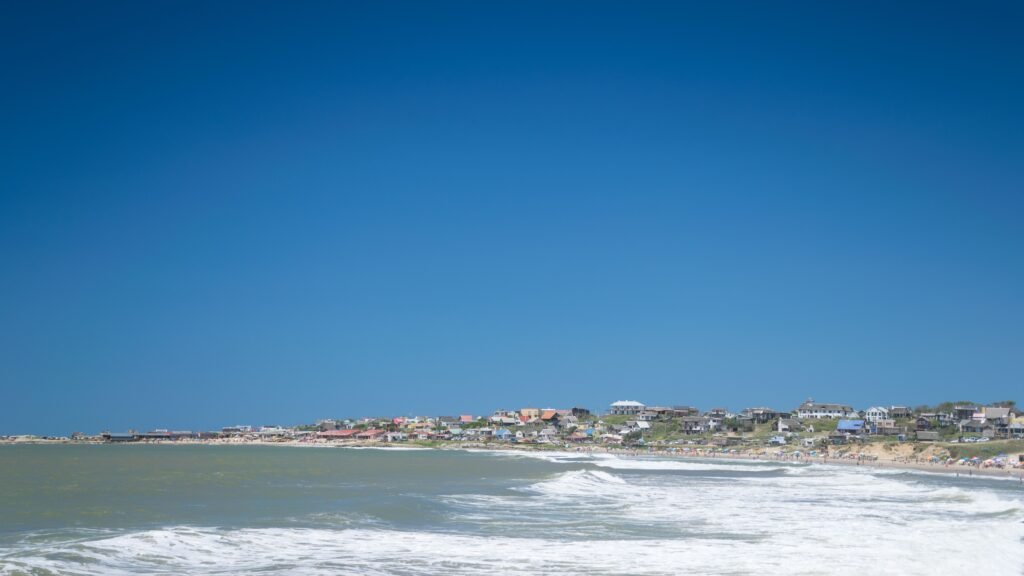 Cidade litorânea com praia de areia, casas coloridas e pessoas perto da costa sob um céu azul claro.