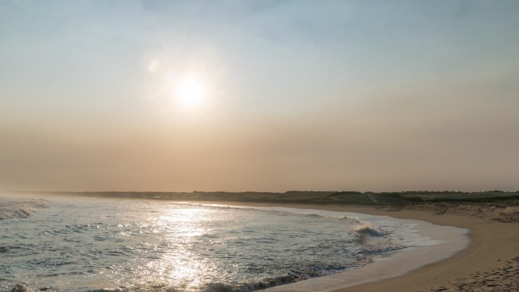 Pôr do sol sobre uma praia tranquila com ondas suaves e dunas de areia clara no horizonte.