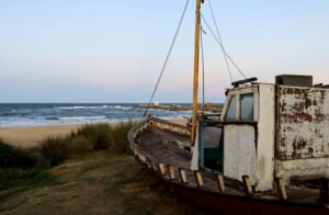 Barco enferrujado e abandonado em uma duna gramada perto da praia, com o mar ao fundo.