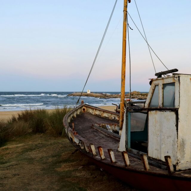 Barco enferrujado e abandonado em uma duna gramada perto da praia, com o mar ao fundo.