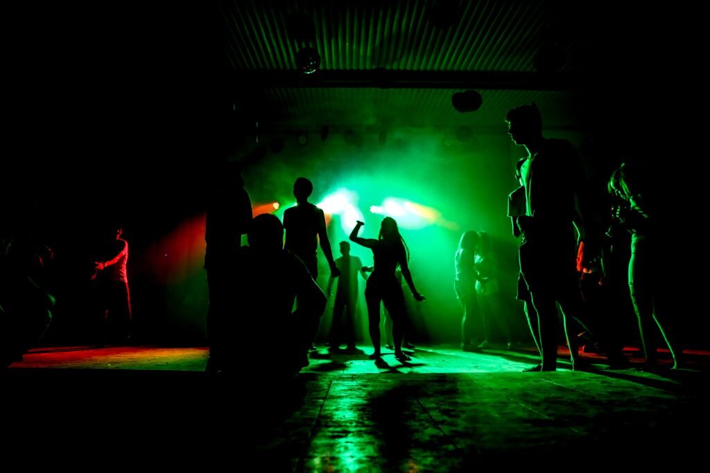 Silhuetas de uma multidão dançando em uma boate escura, com iluminação verde e nebulosa e holofotes acima.
