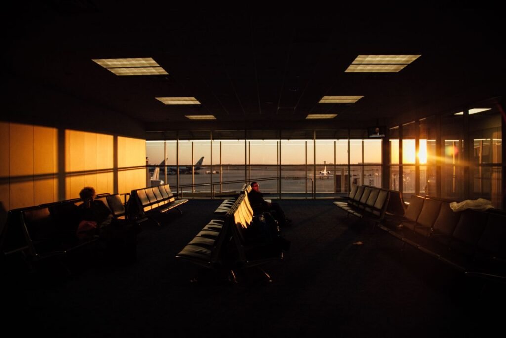 Sunset sunlight fills an empty airport terminal's seating area, with a lone traveler and planes visible through large windows outside.