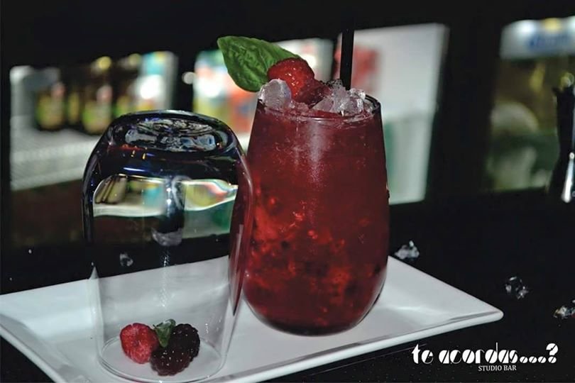 Coquetel vermelho com gelo e hortelã como guarnição em um prato branco ao lado de um copo de água em um bar.