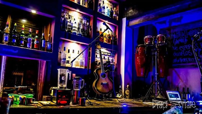 Palco de bar com um violão acústico em um suporte, congas, microfone e um laptop; prateleiras de garrafas brilham em luzes azuis e roxas ao fundo.