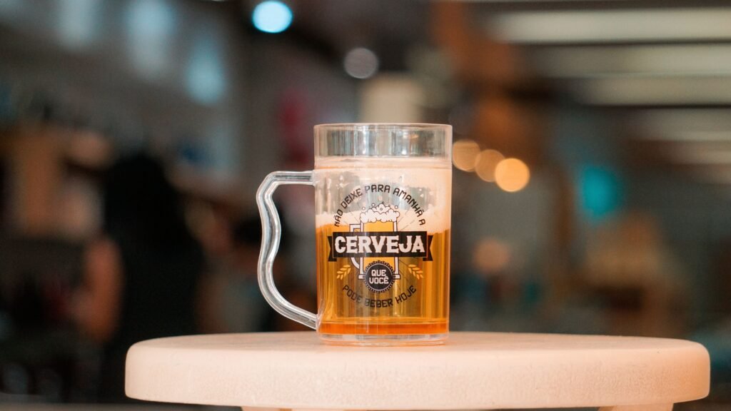 Caneca alta de cerveja com líquido âmbar e espuma cremosa sobre um balcão branco, com fundo desfocado de um bar.