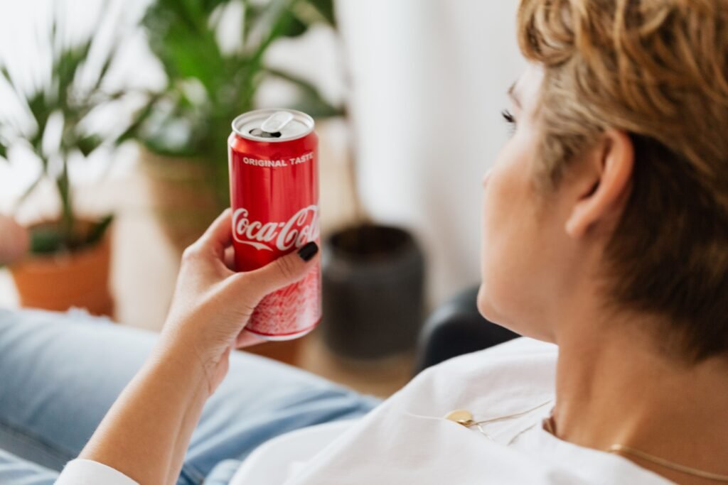 Pessoa entregando uma lata vermelha de Coca-Cola para outra pessoa em ambiente interno; close-up da lata e das mãos.