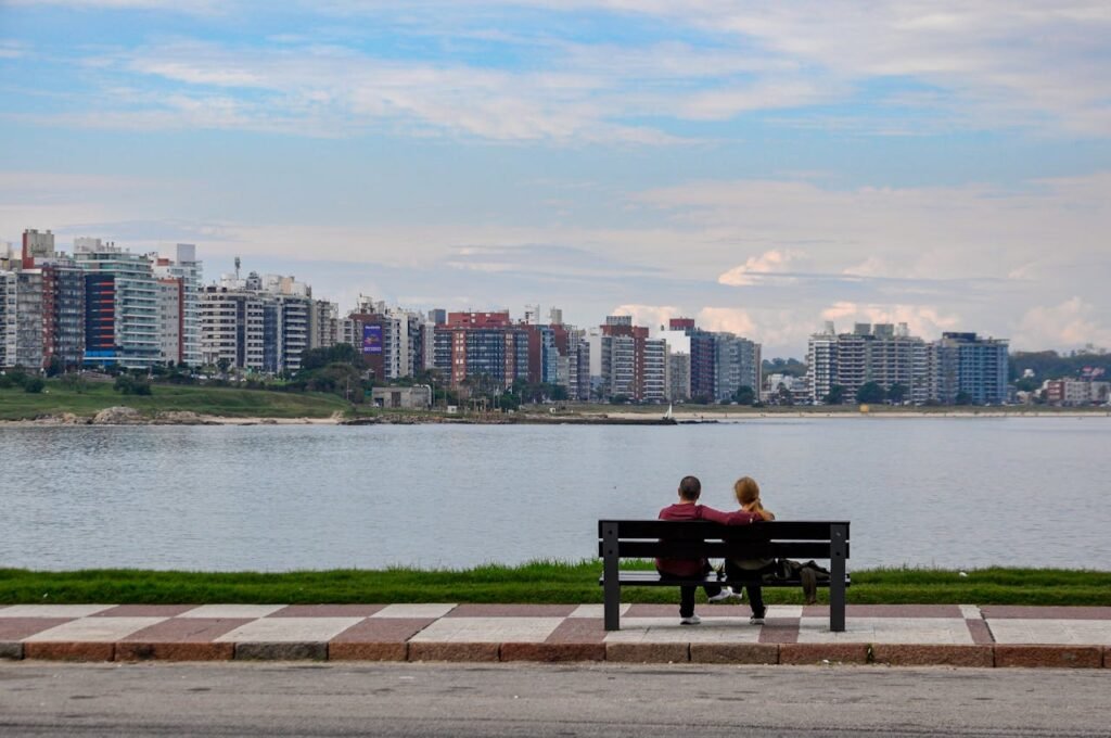 Couple sitting on a bench by the water with a city skyline in the background.