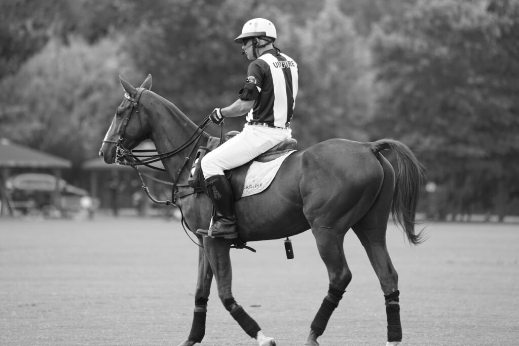 Árbitro de polo a cavalo, usando capacete e uma camisa listrada com a palavra "ÁRBITRO" nas costas, cavalgando em um campo casino no Montevideo Shopping