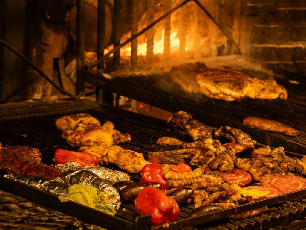 Diversos alimentos grelhados em uma churrasqueira, incluindo pimentões, frango e itens embrulhados em papel alumínio, com uma fogueira aconchegante ao fundo casino no Montevideo Shopping