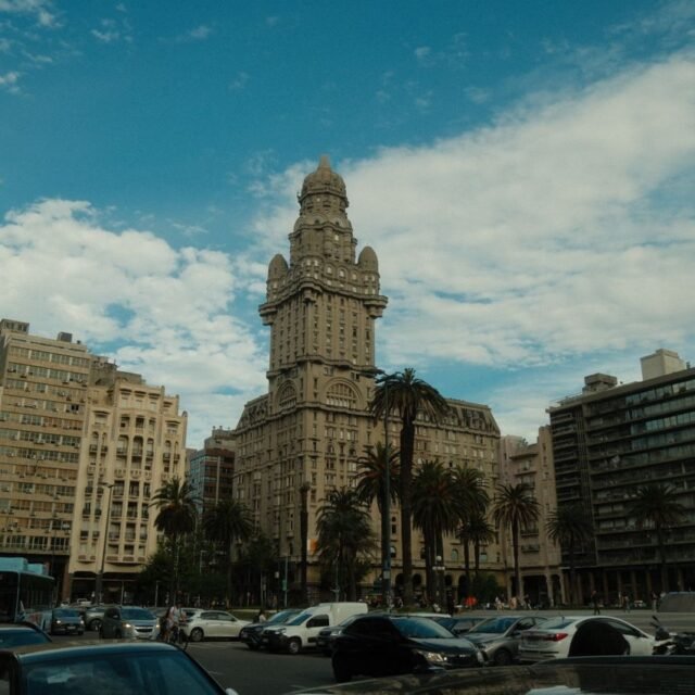 Tall ornate towered building in a busy city square, flanked by palm trees and mid-rise buildings under a blue sky with scattered clouds, cars and pedestrians in the foreground.