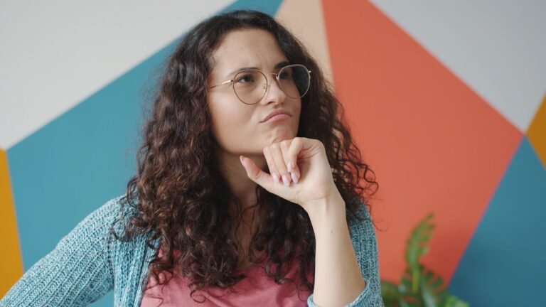 Woman with curly hair and glasses resting her chin thoughtfully in front of a colorful geometric wall.