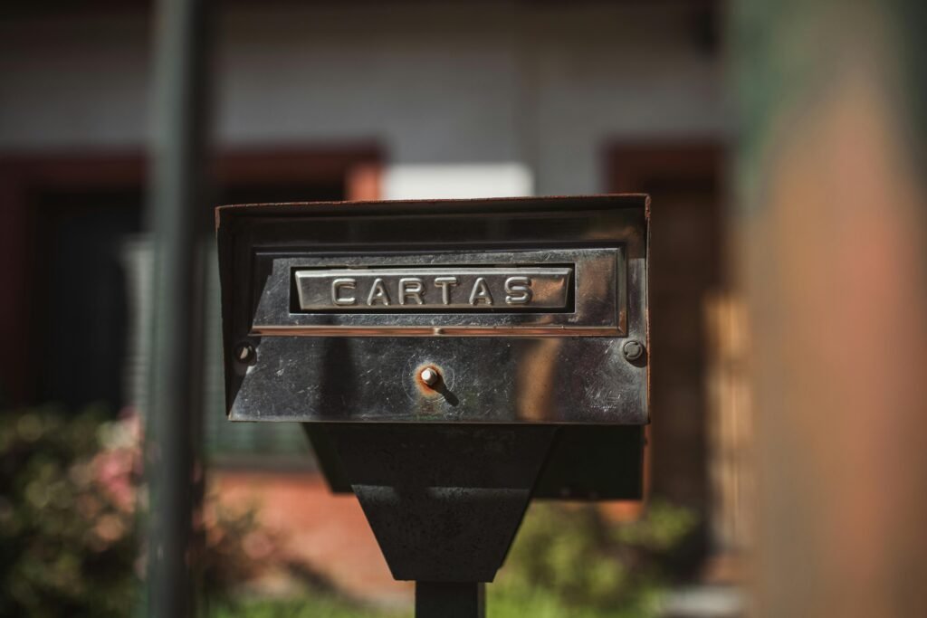 Caixa de correio de metal enferrujado com abertura frontal, fixada em um poste em um ambiente externo ensolarado. Ela está desgastada e em foco no primeiro plano, com uma casa e vegetação desfocadas ao fundo correios no Uruguai