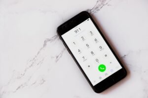 Visão aérea de um smartphone preto deitado na diagonal sobre uma superfície de mármore branco com veios cinzas. A tela do aparelho está ativa, exibindo o aplicativo de discagem telefônica com o número de emergência "911" digitado no topo.