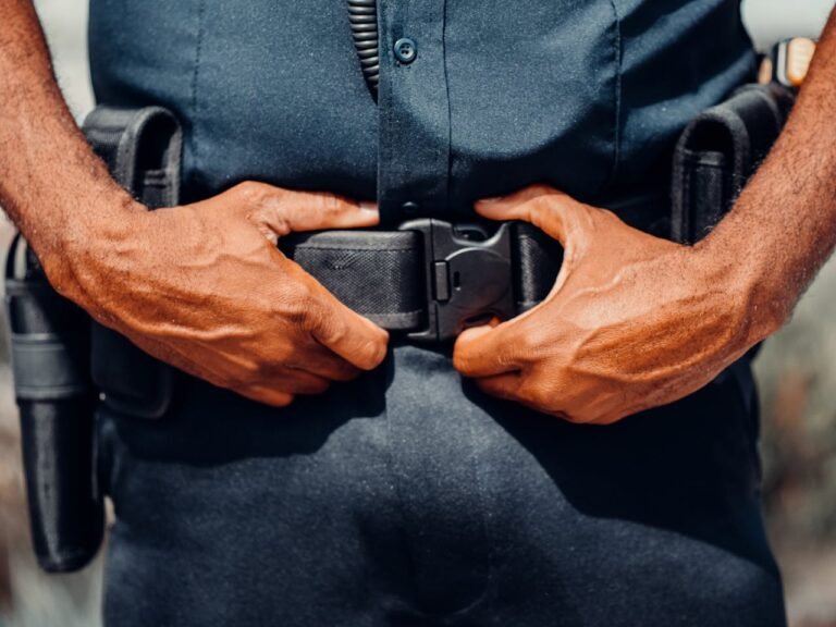 Foto em primeiro plano focada na cintura de um oficial de uniforme azul-escuro ajustando a fivela plástica de seu cinto tático de utilidades. A imagem, que destaca os equipamentos de segurança pública e policiamento urbano, serve como ilustração para o tema de delegacias de policia em montevideo.