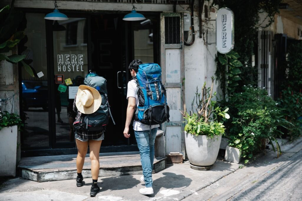 Dois mochileiros com mochilas grandes se aproximam da entrada de um albergue com a placa 'HERE HOSTEL' em uma rua da cidade.