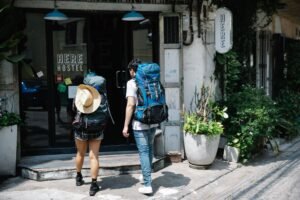 Dois mochileiros com mochilas grandes se aproximam da entrada de um albergue com a placa 'HERE HOSTEL' em uma rua da cidade.