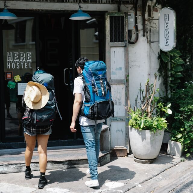Dois mochileiros com mochilas grandes se aproximam da entrada de um albergue com a placa 'HERE HOSTEL' em uma rua da cidade.